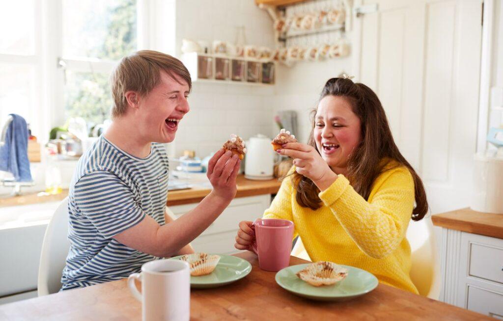 Two people laughing and sharing cupcakes at a kitchen table, representing NDIS daily living support on the Gold Coast.